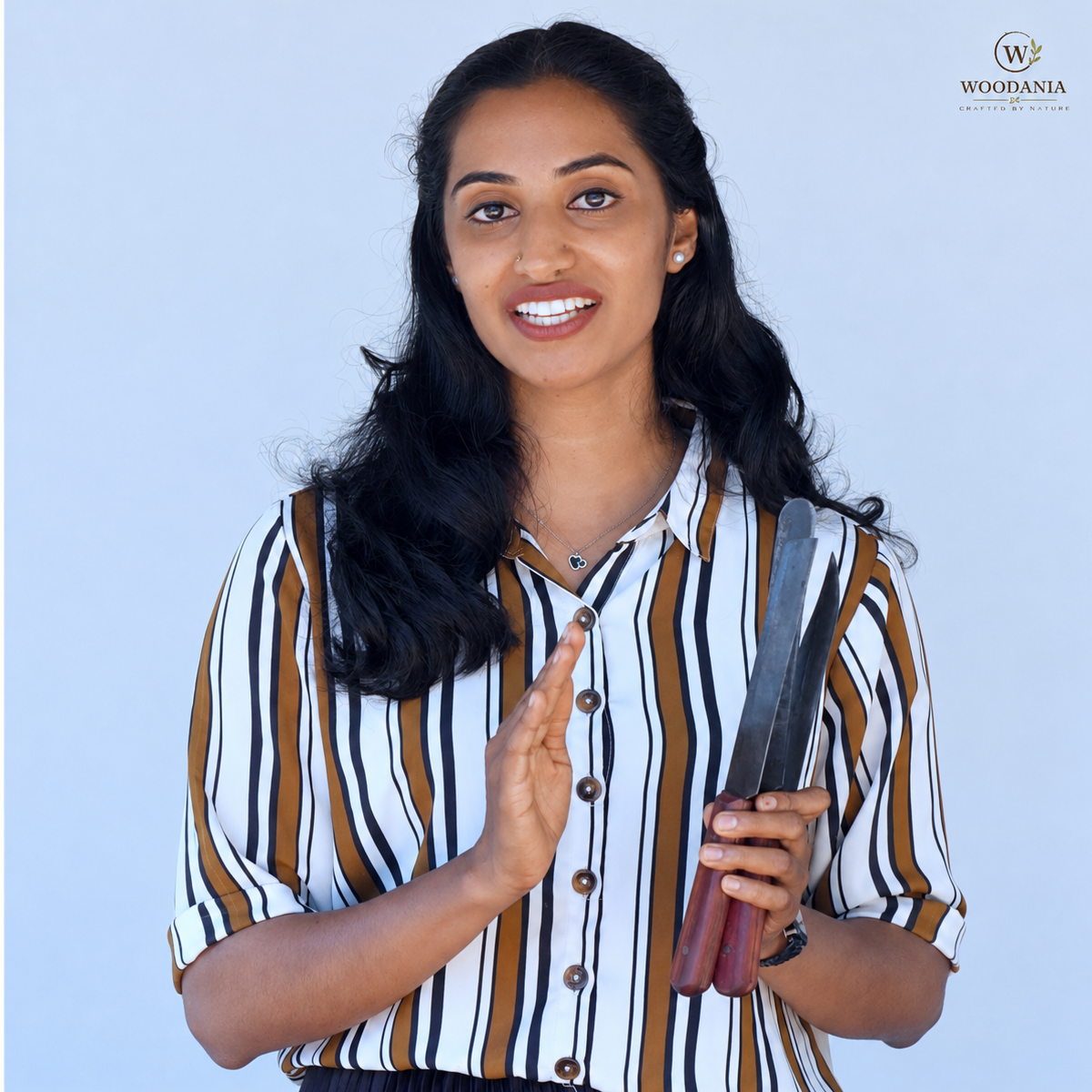 A smiling woman with long dark hair stands against a light blue background, wearing a white button-up shirt with vertical brown and black stripes. She holds a pair of metal knives with reddish-brown handles.